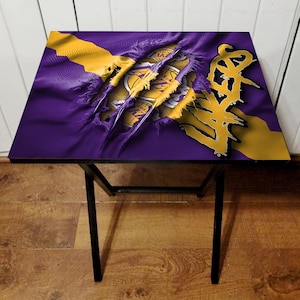May include: A black folding table with a purple and gold Lakers themed design. The design features a ripped fabric effect with the Lakers logo and the word "LAKERS" in a bold font.