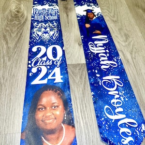 May include: Blue and white graduation stole with a white background and a blue and white splatter pattern. The stole has the name "Myiah Broyles" and the text "Class of 2024" on one side. The other side has a white tiger mascot, the text "Fayette-Ware High School", and a photo of a graduate.