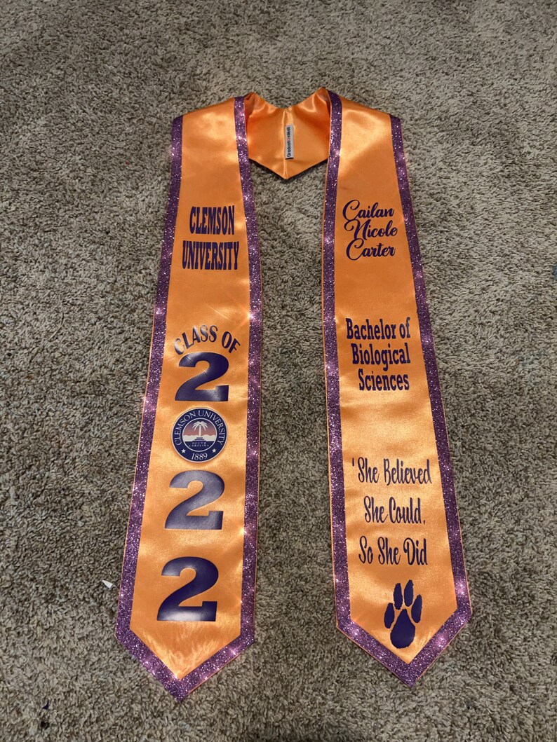 May include: Orange and purple graduation stole with the text "Clemson University", "Class of 2022", "Cailan Nicole Carter", "Bachelor of Biological Sciences", and "She Believed She Could, So She Did".