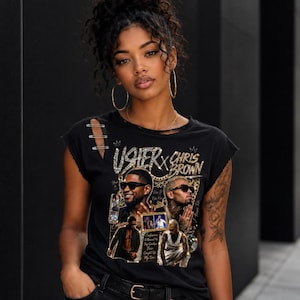 May include: A black short-sleeve t-shirt featuring a graphic print of Usher and Chris Brown with gold chain accents and text. The shirt has a distressed neckline and shoulder detail with three silver safety pins attached to the right shoulder area.