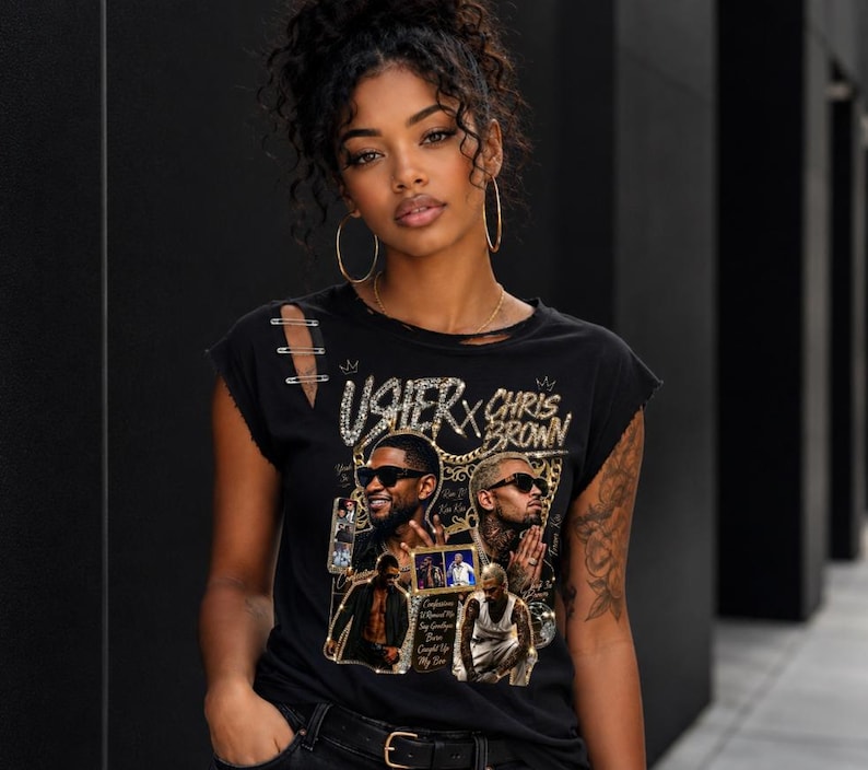May include: A black short-sleeve t-shirt featuring a graphic print of Usher and Chris Brown with gold chain accents and text. The shirt has a distressed neckline and shoulder detail with three silver safety pins attached to the right shoulder area.