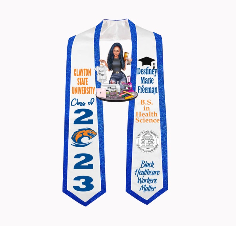 May include: A white graduation sash with blue trim and a blue and orange logo. The sash has the text "Clayton State University" and "Class of 2023". The sash also has the text "Destiney Marie Freeman, B.S. in Health Science" and "Black Healthcare Workers Matter".