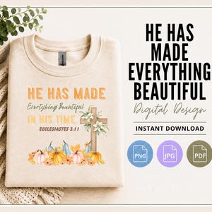 May include: A beige crewneck sweatshirt featuring a graphic print with the text "HE HAS MADE EVERYTHING BEAUTIFUL IN HIS TIME ECCLESIASTES 3:11" alongside a wooden cross, white flowers, orange and blue pumpkins, and autumn leaves.