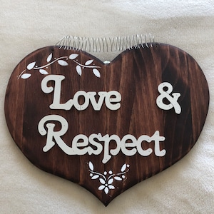 May include: A heart-shaped wooden sign with the words "Love & Respect" in white lettering. The dark brown wood is decorated with white painted floral accents and a silver wire coil at the top.