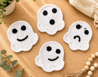 May include: Four white crocheted ghost coasters with black yarn details. Each ghost features a unique facial expression: one smiling, one with a surprised O-shaped mouth, one frowning, and one with a tilted smile. The coasters are flat and circular.