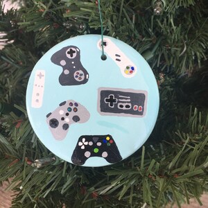 May include: A light blue Christmas ornament with a painted design of various video game controllers, including a joystick, a game pad, and a classic NES controller.