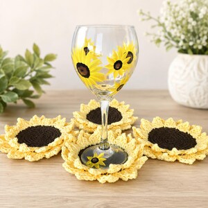 May include: A clear glass wine glass painted with yellow and brown sunflower designs, accompanied by four round, crocheted sunflower coasters featuring dark brown centers and bright yellow petals arranged in a circular pattern.