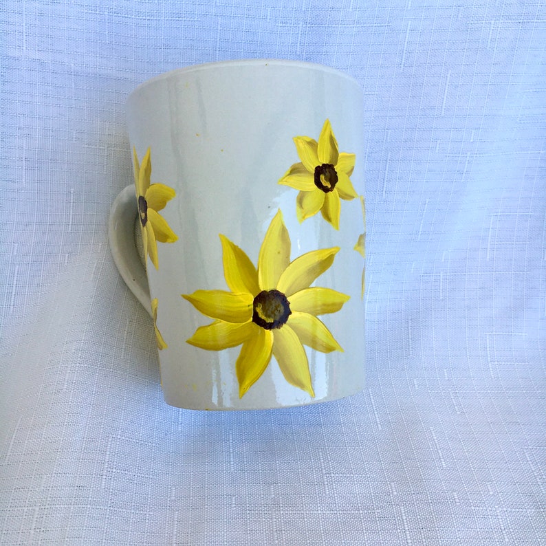 K&ouml;nnte beinhalten: Wei&szlig;e Keramiktasse mit handbemaltem Design von gelben Sonnenblumen. Die Tasse hat einen abgerundeten Griff.