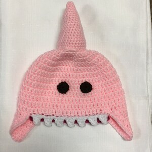 May include: Pink crocheted shark hat with black eyes and white teeth. The hat has a pointed fin on top.