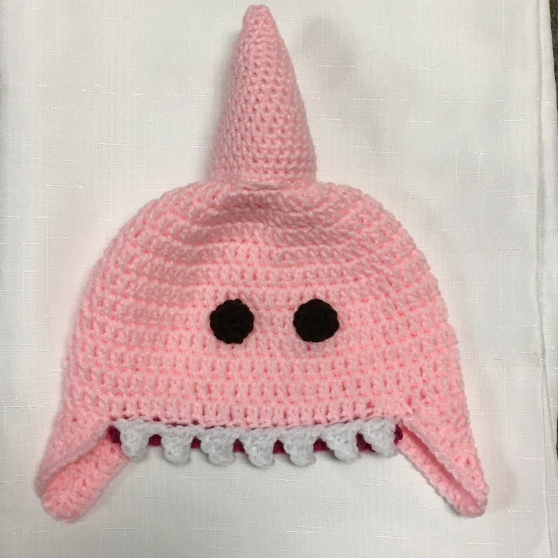 May include: Pink crocheted shark hat with black eyes and white teeth. The hat has a pointed fin on top.