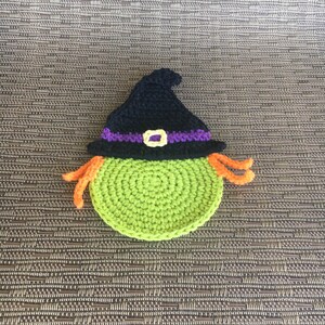 Op de afbeelding: Groene gehaakte heks met een zwarte hoed en oranje haar. De heks draagt een paarse en gele band om haar hoed.