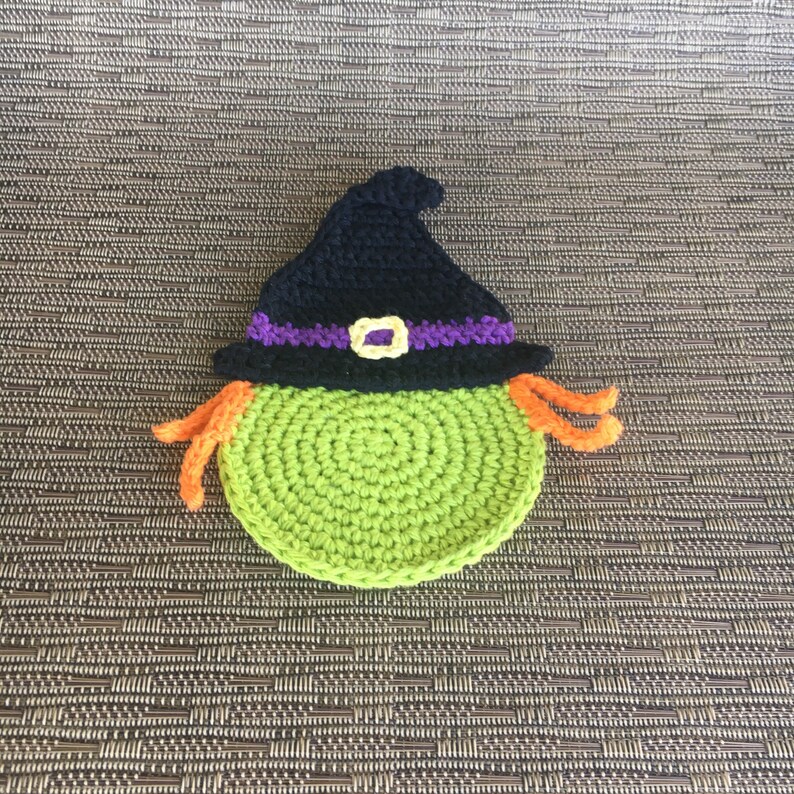 Op de afbeelding: Groene gehaakte heks met een zwarte hoed en oranje haar. De heks draagt een paarse en gele band om haar hoed.
