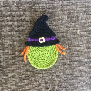 Op de afbeelding: Een groene en zwarte gebreide heks met oranje haar. De heks draagt een zwarte hoed met een paarse band en een gouden gesp.