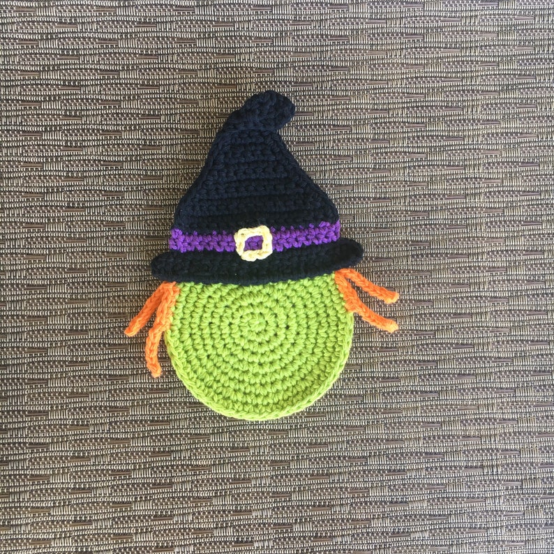 Op de afbeelding: Een groene en zwarte gebreide heks met oranje haar. De heks draagt een zwarte hoed met een paarse band en een gouden gesp.