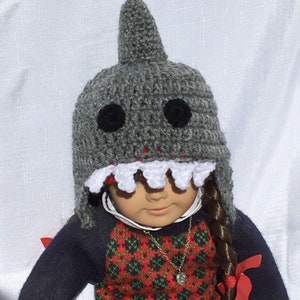 May include: A gray crocheted shark hat with white teeth and black eyes. The hat is worn by a doll with long brown hair and a red and green plaid sweater.