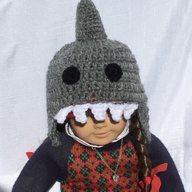 May include: A gray crocheted shark hat with white teeth and black eyes. The hat is worn by a doll with long brown hair and a red and green plaid sweater.