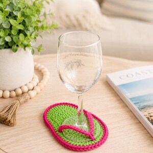 May include: A clear glass wine glass with the etched text New Hope Wine and a botanical illustration, resting on a bright green and hot pink crocheted coaster shaped like a flip-flop sandal with a pink strap detail across the top surface.