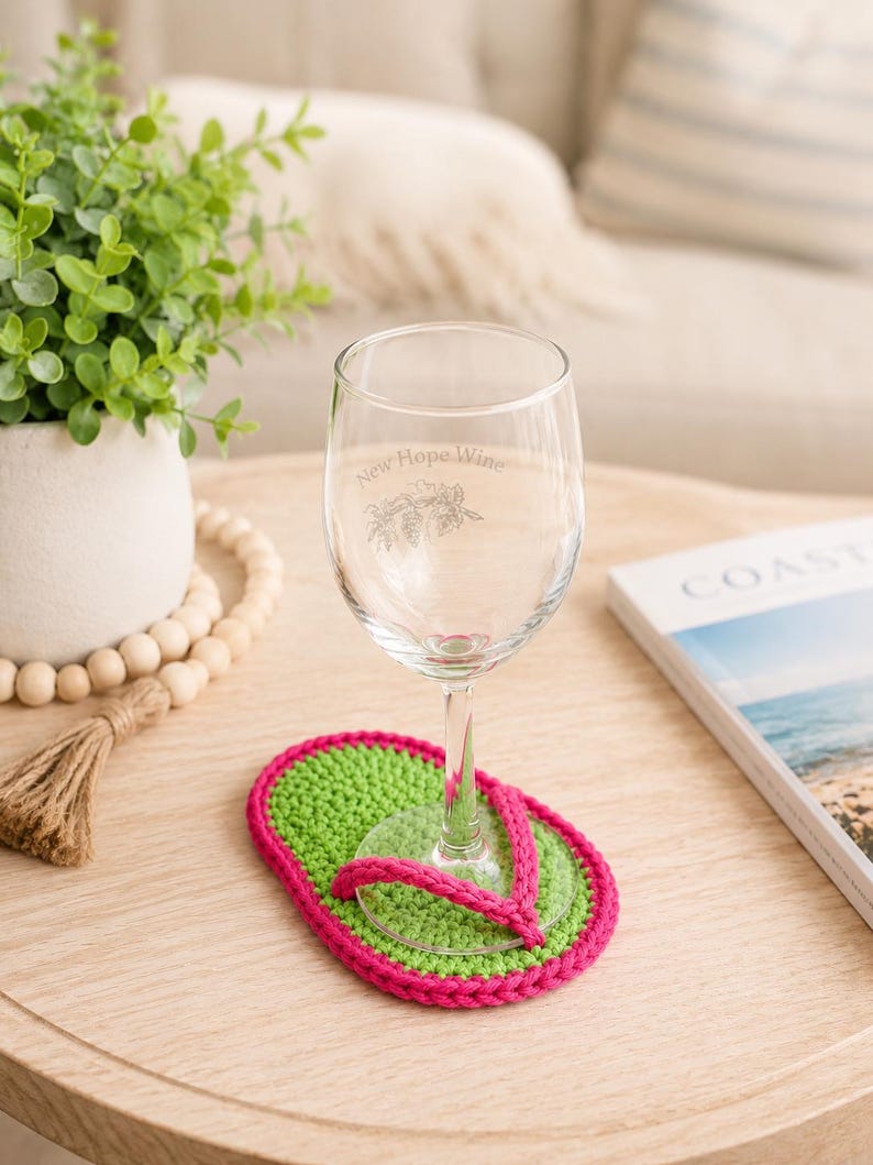 May include: A clear glass wine glass with the etched text New Hope Wine and a botanical illustration, resting on a bright green and hot pink crocheted coaster shaped like a flip-flop sandal with a pink strap detail across the top surface.