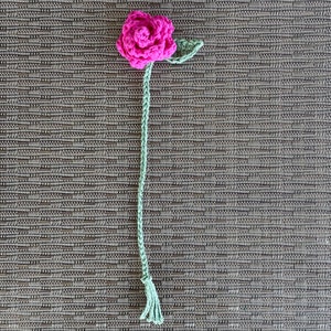 以下が含まれることがあります： 緑色の茎と葉が付いた、ピンク色の毛糸で編まれたバラ。バラは、長い緑色の毛糸の房に繋がっています。