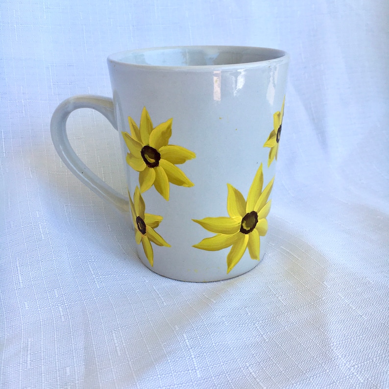 K&ouml;nnte beinhalten: Wei&szlig;e Keramiktasse mit handbemaltem Design aus gelben Blumen mit braunen Zentren. Die Blumen sind um die Tasse herum verstreut.