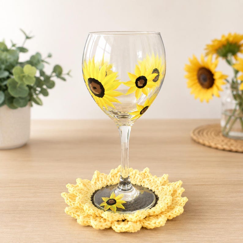 May include: A clear glass wine glass featuring hand-painted yellow sunflower designs with dark brown centers, resting on a round, yellow crocheted sunflower-shaped coaster with a textured petal edge and a small matching sunflower motif.