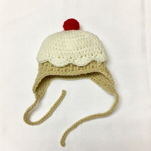 May include: A crocheted ice cream cone hat with a red cherry on top. The hat is beige and white with a long beige tie.