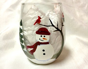 May include: Hand-painted wine glass featuring a snowman with a red hat and scarf, a cardinal perched on a branch, and a green pine tree. The glass is filled with white tissue paper.