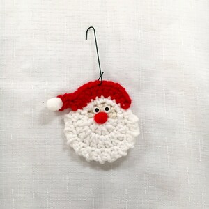 Peut inclure: Ornement de No&euml;l en crochet blanc repr&eacute;sentant le P&egrave;re No&euml;l avec un chapeau rouge et un pompon blanc. L'ornement a des yeux noirs et un nez rouge.