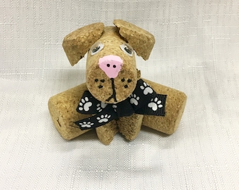 May include: A brown cork dog with black and white paw print ribbon, white googly eyes, and a pink nose.