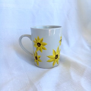 K&ouml;nnte beinhalten: Wei&szlig;e Keramiktasse mit gelbem und braunem Blumendesign. Die Tasse hat einen Henkel und ist mit vier gelben Bl&uuml;ten mit braunen Zentren verziert.