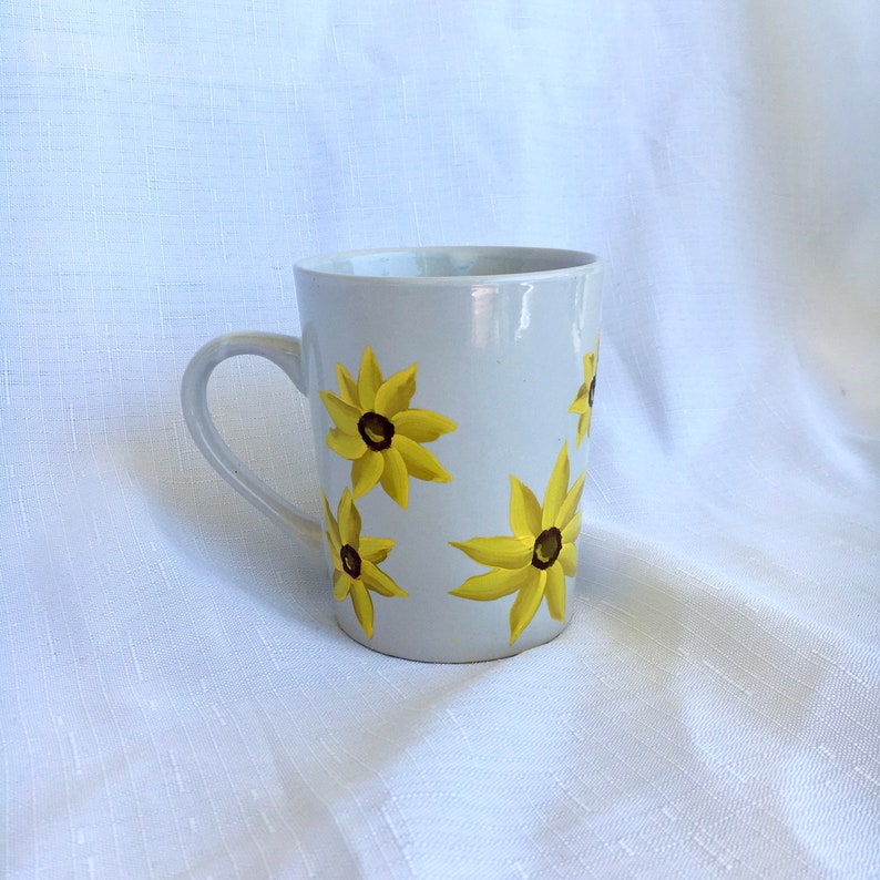 K&ouml;nnte beinhalten: Wei&szlig;e Keramiktasse mit gelbem und braunem Blumendesign. Die Tasse hat einen Henkel und ist mit vier gelben Bl&uuml;ten mit braunen Zentren verziert.
