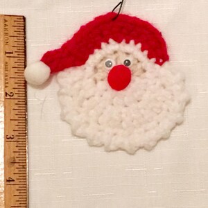 Peut inclure: Une d&eacute;coration de No&euml;l en crochet repr&eacute;sentant un P&egrave;re No&euml;l avec une barbe blanche, un chapeau rouge et des yeux noirs. La d&eacute;coration est suspendue &agrave; une ficelle.
