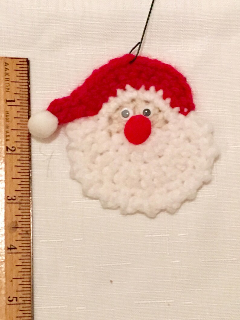 Peut inclure: Une d&eacute;coration de No&euml;l en crochet repr&eacute;sentant un P&egrave;re No&euml;l avec une barbe blanche, un chapeau rouge et des yeux noirs. La d&eacute;coration est suspendue &agrave; une ficelle.