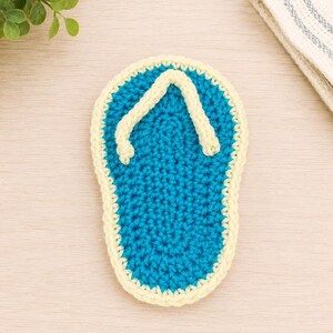 May include: A crochet flip-flop coaster featuring a bright blue sole with a textured stitch pattern and a pale yellow border. The thong strap is also crafted in pale yellow yarn, creating a distinct outline against the blue center of the piece.