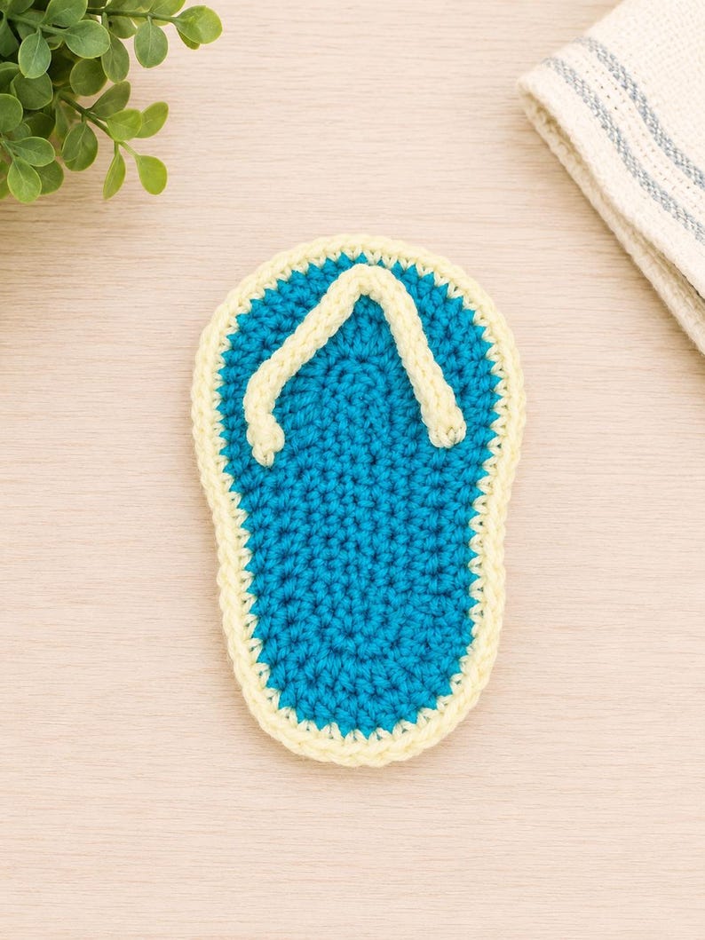May include: A crochet flip-flop coaster featuring a bright blue sole with a textured stitch pattern and a pale yellow border. The thong strap is also crafted in pale yellow yarn, creating a distinct outline against the blue center of the piece.