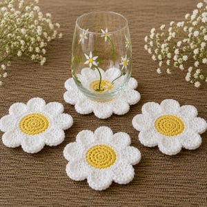 May include: A clear glass decorated with daisy flowers rests on a white crochet daisy coaster. Three more white crochet daisy coasters with yellow centres are arranged around the glass. The coasters are on a brown surface.