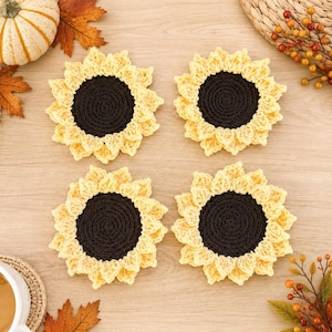 May include: Four round crochet coasters designed to resemble sunflowers, each featuring a dark brown circular center surrounded by light yellow petals. The coasters are arranged in a square formation, showcasing the detailed yarn texture and stitching.