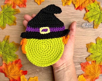 May include: A round, lime green crocheted coaster featuring a black witch hat with a purple band and a yellow buckle. The coaster has small orange yarn accents on the sides, representing hair, and is crafted with a dense, circular stitch pattern.