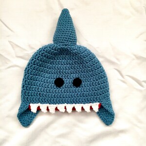 May include: A blue crocheted shark hat with black eyes and white teeth. The hat has a pointed fin on top and two floppy ears.