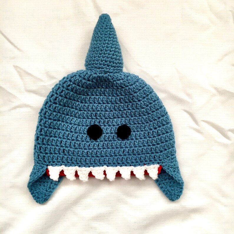 May include: A blue crocheted shark hat with black eyes and white teeth. The hat has a pointed fin on top and two floppy ears.