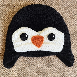 May include: A black and white crochet hat with a penguin face design. The hat features a black body, white face, black eyes, and an orange beak. The hat has ear flaps.