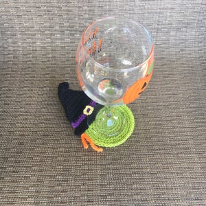 Op de afbeelding: Een groene en zwarte gehaakte heksenhoed onderzetter met oranje franjes. De onderzetter bevindt zich onder een helder glas met een geschilderde pompoen en de woorden "Happy Hallowen" erop.