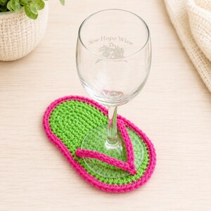 May include: A clear glass wine glass with the etched text New Hope Wine and a floral graphic, resting on a bright green and hot pink crocheted coaster shaped like a flip-flop sandal. The coaster features a vibrant green sole with a pink strap.