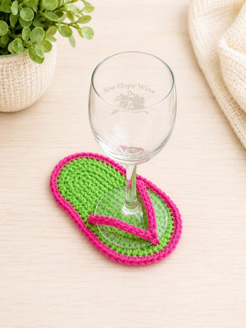 May include: A clear glass wine glass with the etched text New Hope Wine and a floral graphic, resting on a bright green and hot pink crocheted coaster shaped like a flip-flop sandal. The coaster features a vibrant green sole with a pink strap.