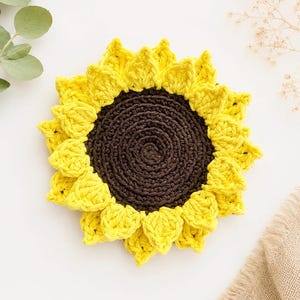 May include: A round, crocheted sunflower coaster featuring a dark brown circular center with a textured, concentric stitch pattern, surrounded by two layers of bright yellow, pointed petals that create a full, dimensional floral appearance.