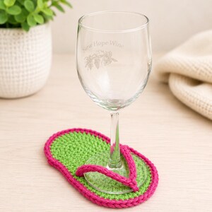 May include: A clear glass wine glass with the etched text New Hope Wine and a grape cluster graphic, resting on a crochet coaster shaped like a flip-flop with a bright green sole and a vibrant pink strap border and toe post.