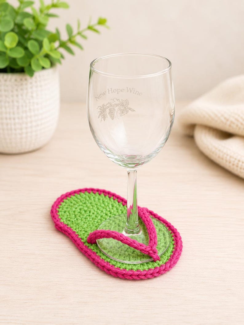 May include: A clear glass wine glass with the etched text New Hope Wine and a grape cluster graphic, resting on a crochet coaster shaped like a flip-flop with a bright green sole and a vibrant pink strap border and toe post.