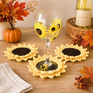 May include: A clear glass wine glass painted with yellow and brown sunflower motifs, accompanied by three round, crocheted sunflower coasters featuring dark brown centers and yellow petals, arranged on a wooden surface.