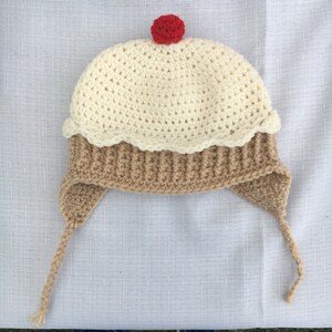 May include: A crocheted hat shaped like a cupcake with a red cherry on top. The hat is white and brown with a scalloped edge.