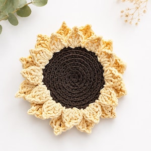 May include: A round, crocheted sunflower coaster featuring a dark brown circular center surrounded by layered, pale yellow petals. The textured yarn construction creates a detailed floral design with a distinct contrast between the dark core and the light outer edge, forming a flat, circular shape.